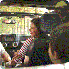 A mother sits in the passenger seat of a vehicle and smiles over her shoulder at her young son in the back seat.
